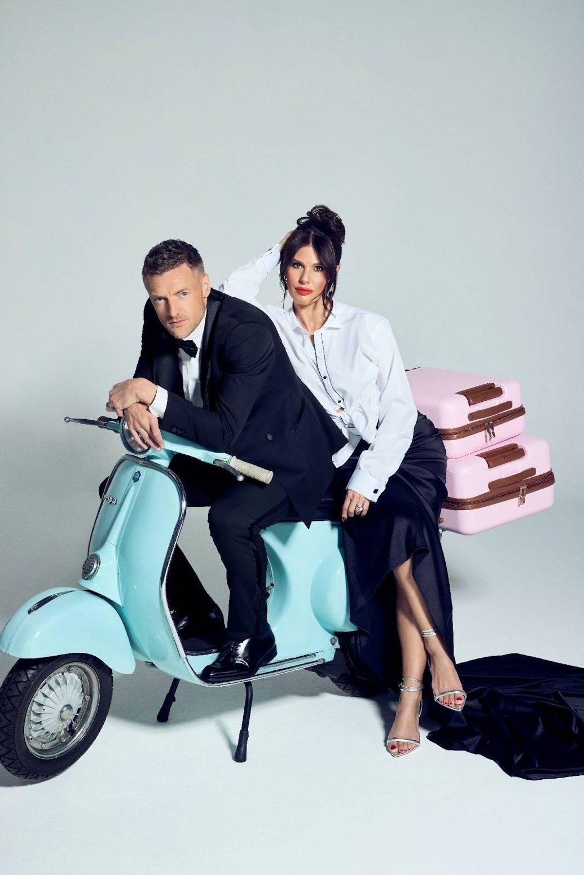 An image of two individuals, dressed formally, seated on a vibrant turquoise scooter, with a pink luggage item in the background, possibly posing for a photograph against a neutral backdrop.