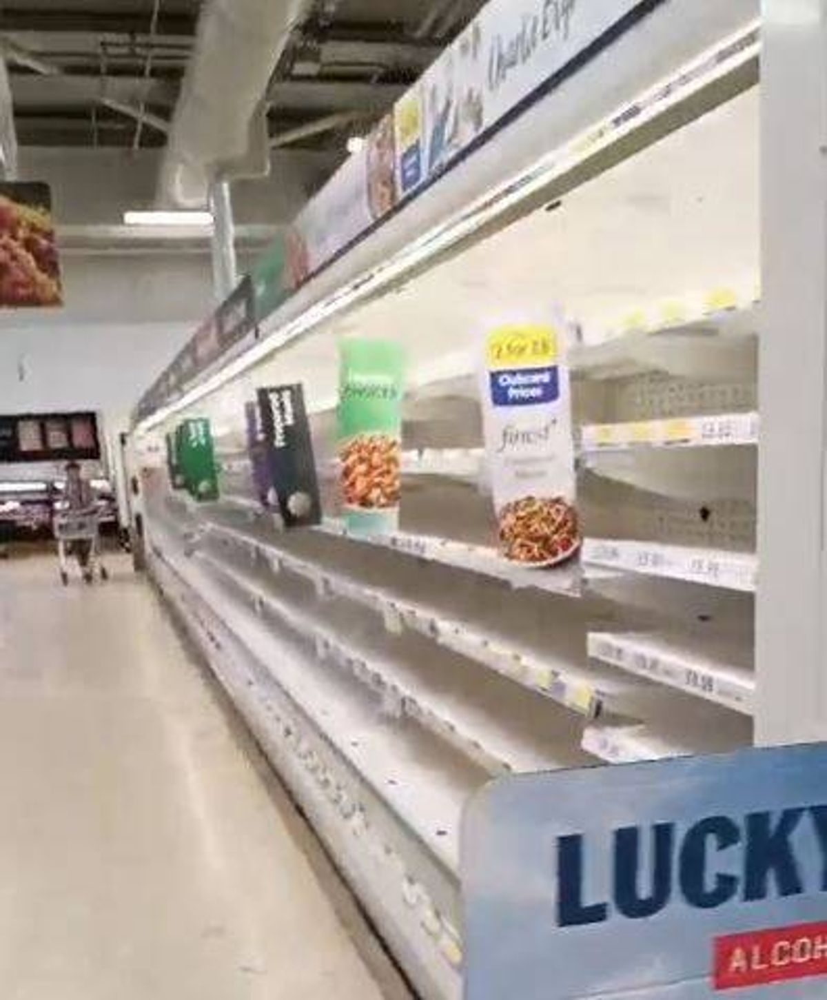An indoor supermarket aisle featuring empty shelves with a few remaining products. A person is walking in the background.
