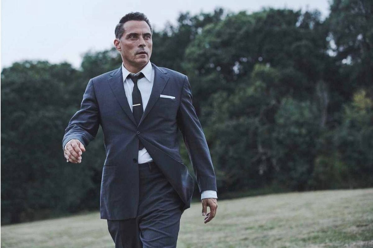 An individual dressed formally in a business suit stands outside, with a background featuring a lush, green landscape. The attire comprises a dark suit jacket, a light-colored dress shirt, a tie, and a crisp white pocket square. The person's expression is contemplative as they gaze forward, suggesting a sense of purpose or deep thought. The natural environment, characterized by a dense collection of trees, provides a serene backdrop that contrasts with the individual's formal appearance.