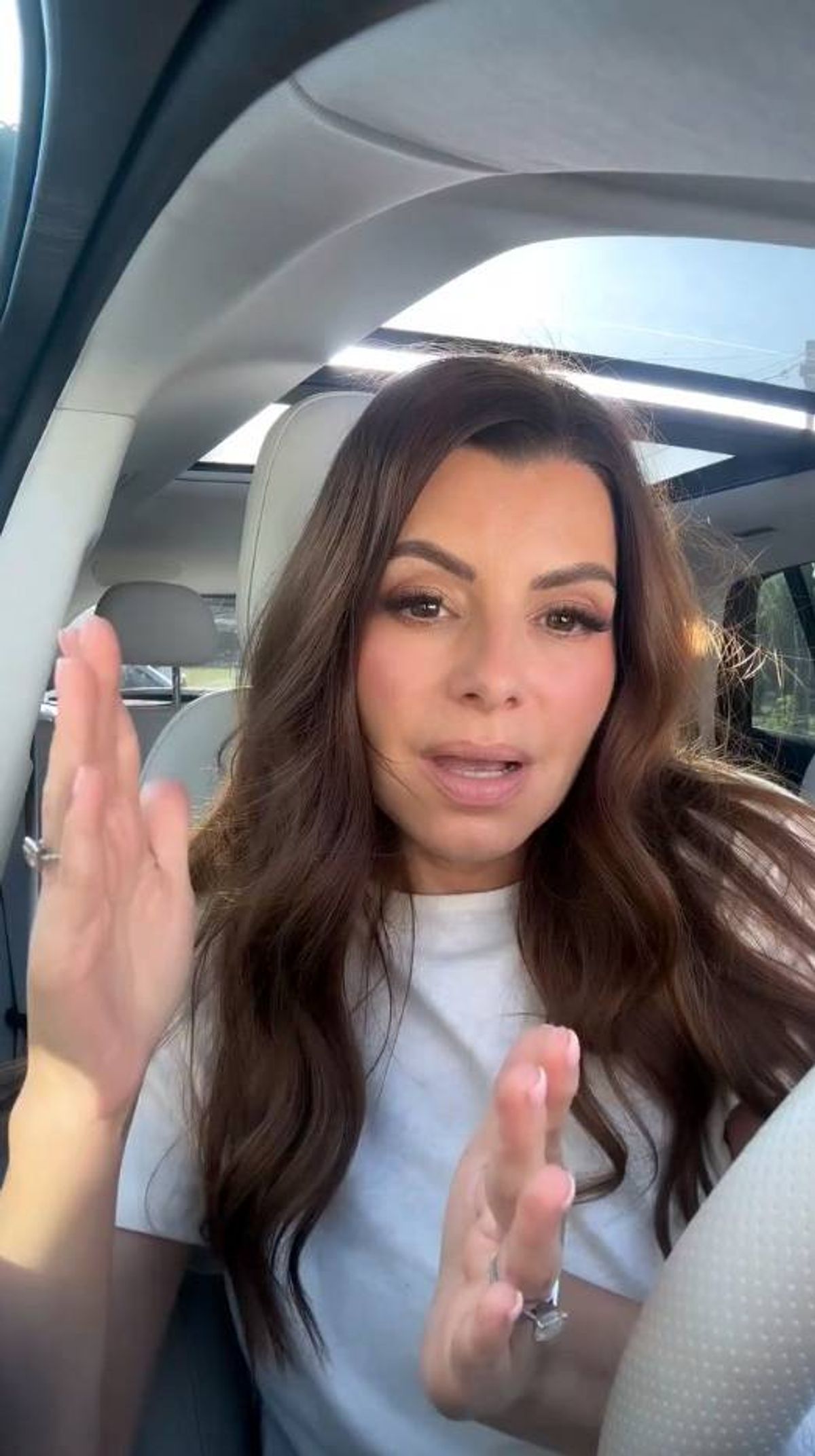 A woman with long, brown hair and a white top is seated inside a vehicle, possibly making a peace sign gesture.