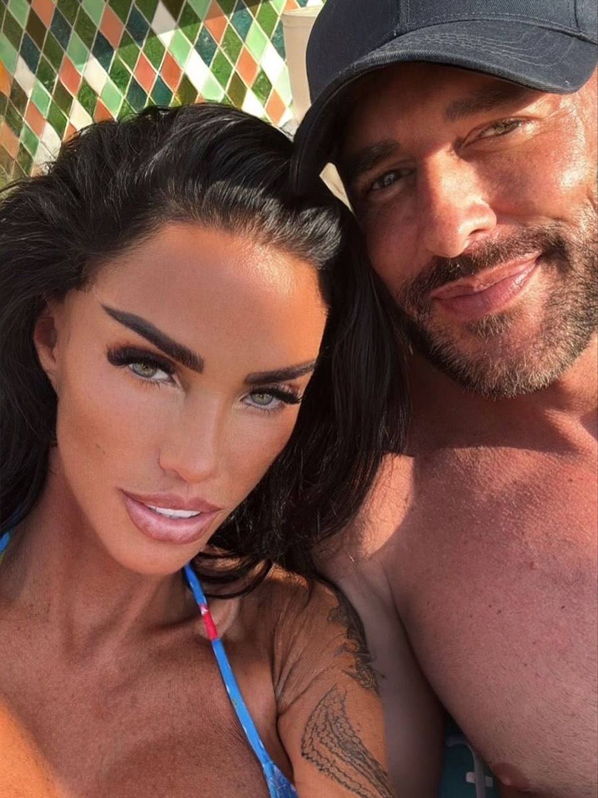 An individual with a striking appearance, featuring prominent eyeliner and a radiant smile, is posing closely beside another individual, who is also smiling and partially revealing his chest.