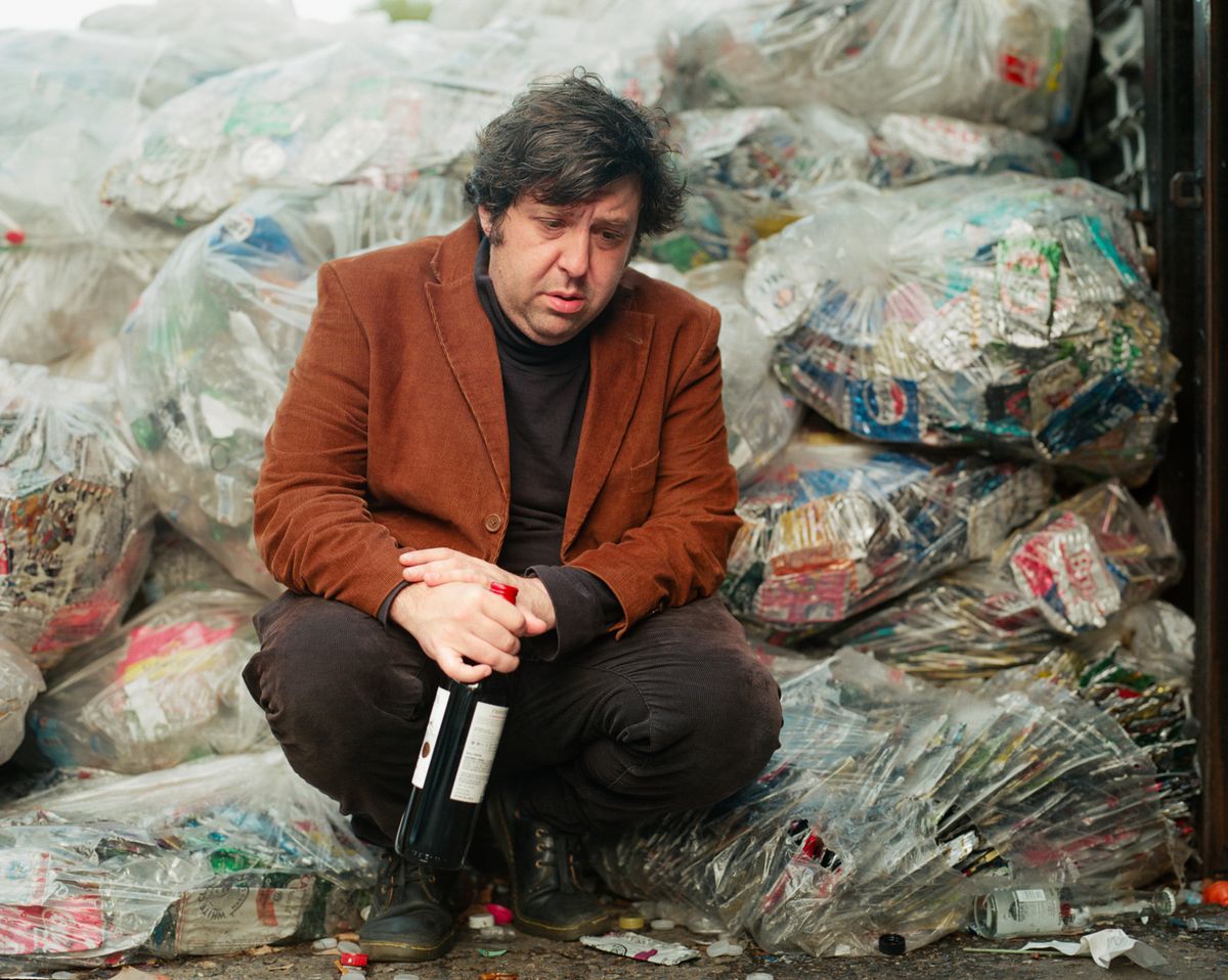 Jim E Brown squats beside a rubbish tip, bottle in hand