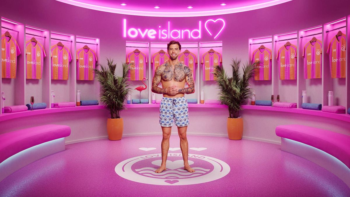 An individual stands in a brightly lit, pink-themed room, possibly a studio, with various decorative elements and vibrant lighting in the background.
