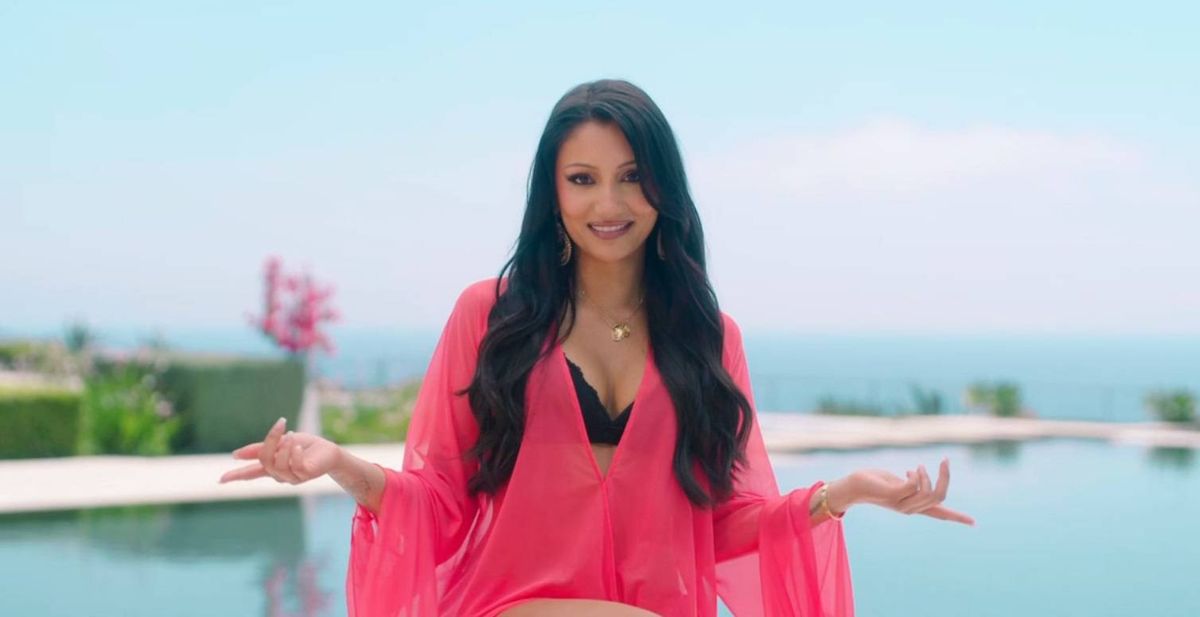 A woman in a pink blouse with long brown hair sits by a pool gesticulating. She is Love Island Series 13 contestant Jasmine Muller.
