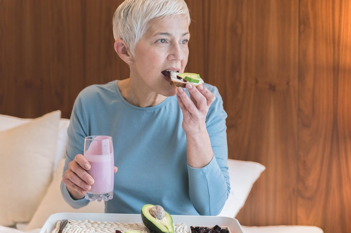 Beautiful senior woman eating nutritious healthy toast with avocado and holding fruit yogurt, Healthy nutrition concept