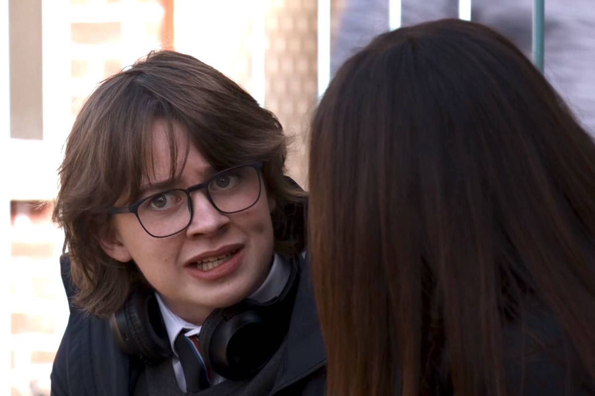 Two individuals are engaged in a conversation. The person on the left is dressed formally, sporting glasses, a necktie, and a headset. The individual on the right has long, dark hair and is dressed casually.