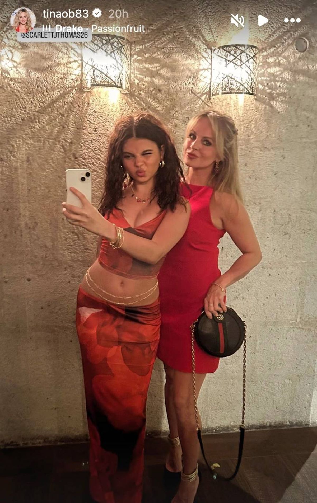 Two individuals, both adorned in red dresses, are captured in a moment of amusement, one of whom is engaged in the act of taking a photograph with a mobile device. The backdrop features a textured wall.