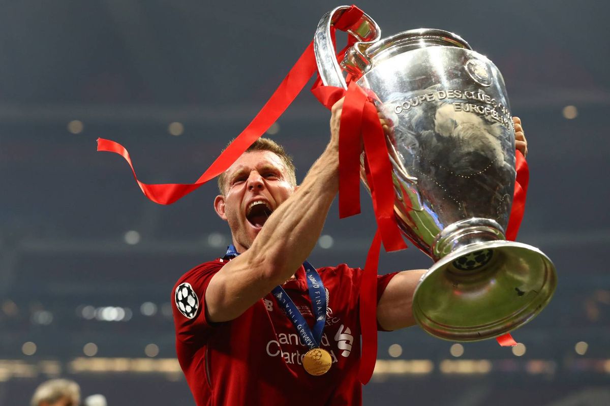 An individual wearing a red jersey, possibly a sports champion, triumphantly raises a large silver trophy adorned with red ribbons above their head in a stadium setting, amidst a backdrop of a cheering crowd.
