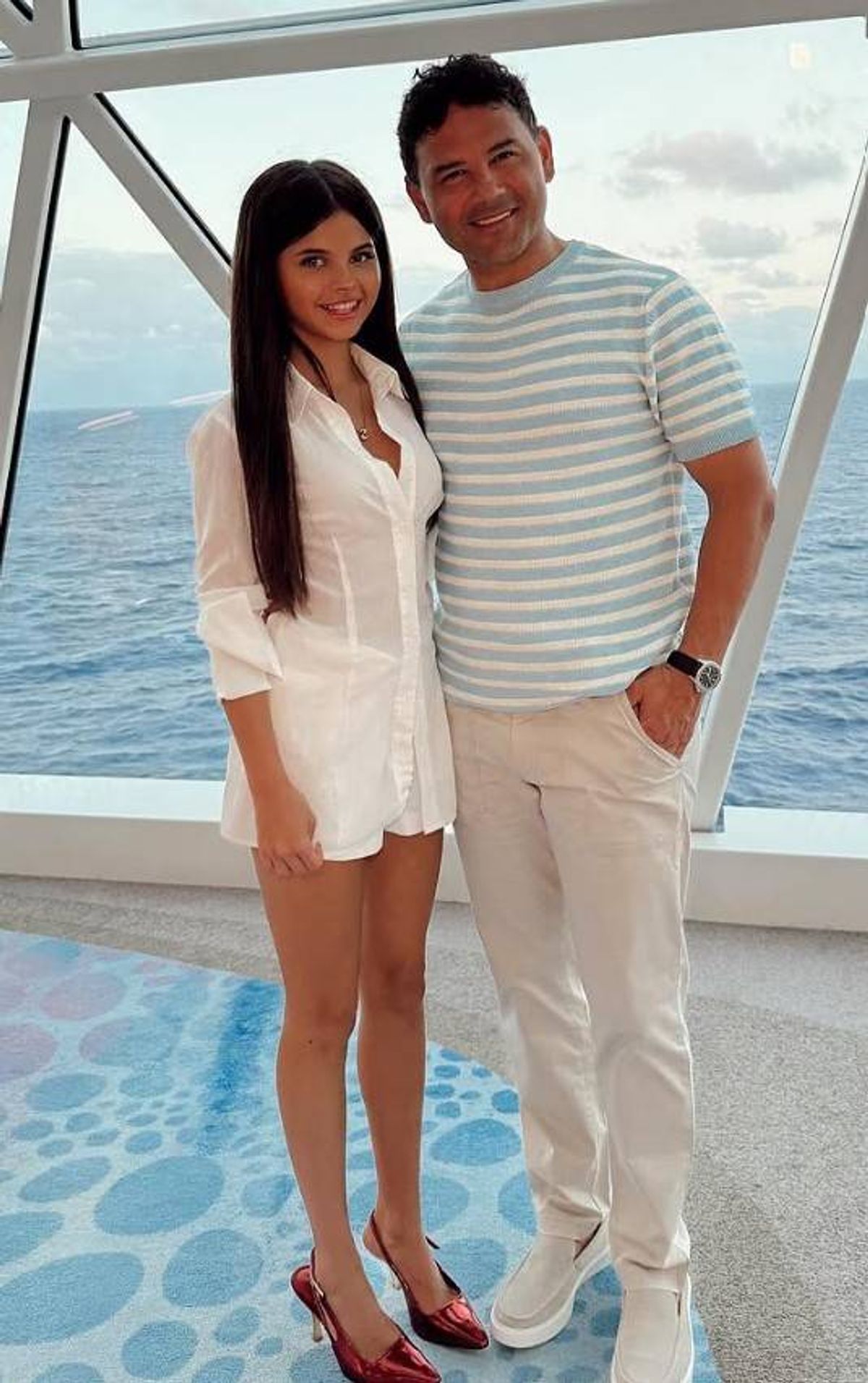An individual dressed in a white shirt and shorts stands next to another person wearing a striped shirt and beige pants, both standing on a deck with an ocean view in the background.