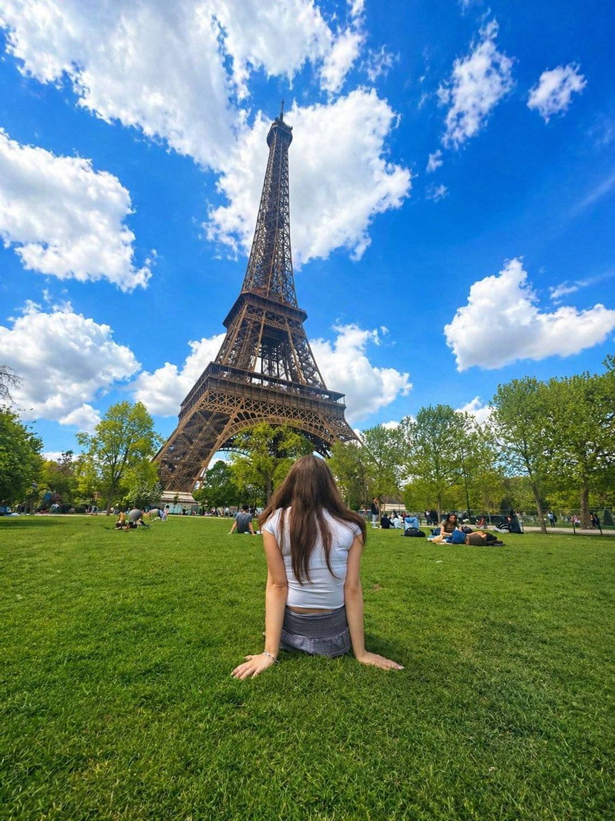A woman is sitting on the grass in front of the Eiffel Tower, surrounded by trees and greenery.