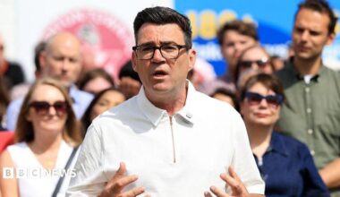 Andy Burnham delivering speech outside in sunny weather in white collared top with zip closure and people behind in sunglasses