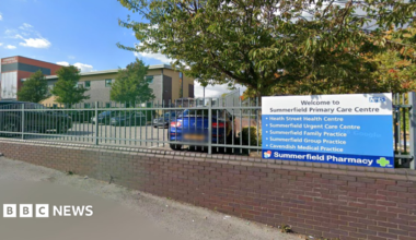 The front of Summerfield Primary Care Centre. There is a blue NHS sign with bullet points on and a sign for the pharmacy. It is on a grey fence and above a brick wall. Behind is a car park and a building.