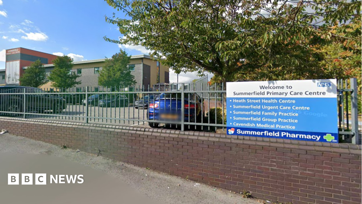 The front of Summerfield Primary Care Centre. There is a blue NHS sign with bullet points on and a sign for the pharmacy. It is on a grey fence and above a brick wall. Behind is a car park and a building.