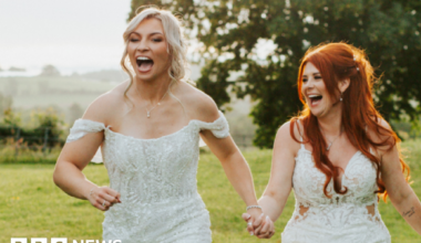 Lauren Price and Carlie Jones walking hand in hand in a field both laughing in white gowns