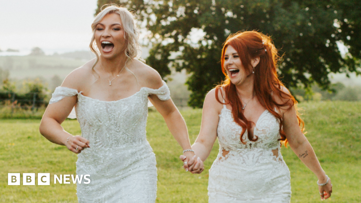 Lauren Price and Carlie Jones walking hand in hand in a field both laughing in white gowns