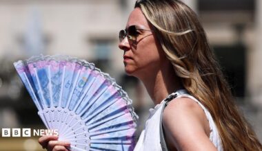 A woman in sunglasses wearing a white top holds a blue and pink fan in London on 24 June 2026