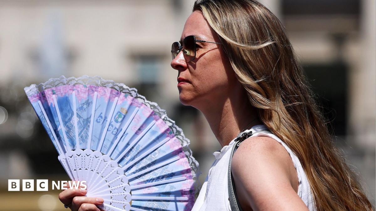 A woman in sunglasses wearing a white top holds a blue and pink fan in London on 24 June 2026