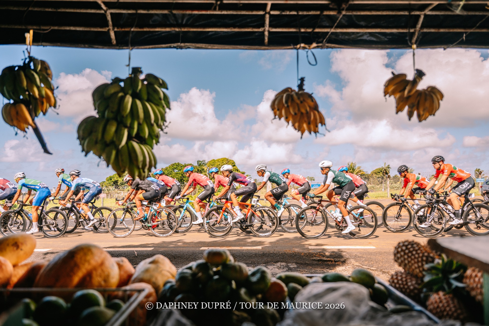 The peloton is pictured from inside a local fruit stand, with various fruits in trays in the foreground and bananas hanging from the awning.