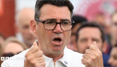 Andy Burnham is captured mid-gesture with both hands raised to chest level, his fingers curled into fists. He is wearing dark-framed glasses and a white zipped top. His mouth is open in mid speech.