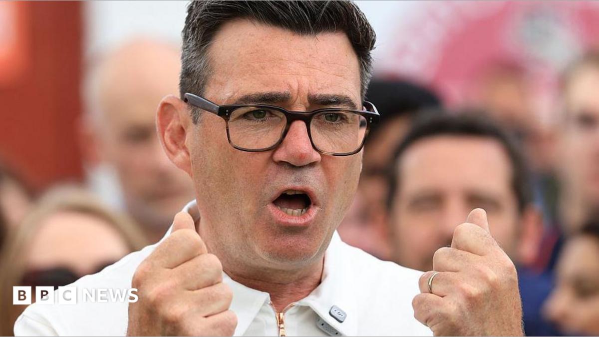 Andy Burnham is captured mid-gesture with both hands raised to chest level, his fingers curled into fists. He is wearing dark-framed glasses and a white zipped top. His mouth is open in mid speech.