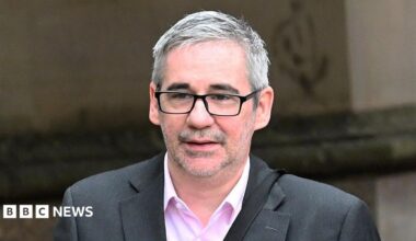 Iain Robertson wears a grey suit and pink shirt. he has short grey hair and wears dark glasses. He is walking outside a building