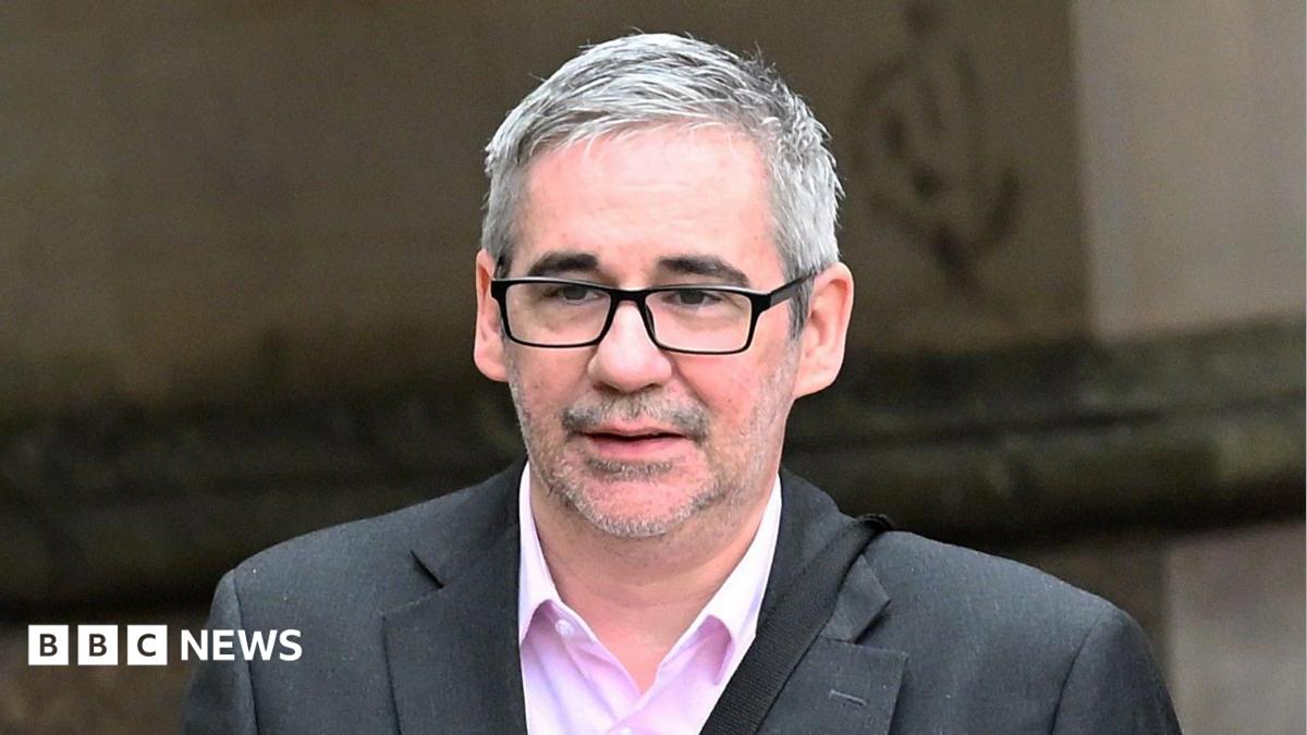 Iain Robertson wears a grey suit and pink shirt. he has short grey hair and wears dark glasses. He is walking outside a building