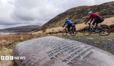 Two people on bikes cycle past a large stone with rugged hills in the background and a loch up ahead