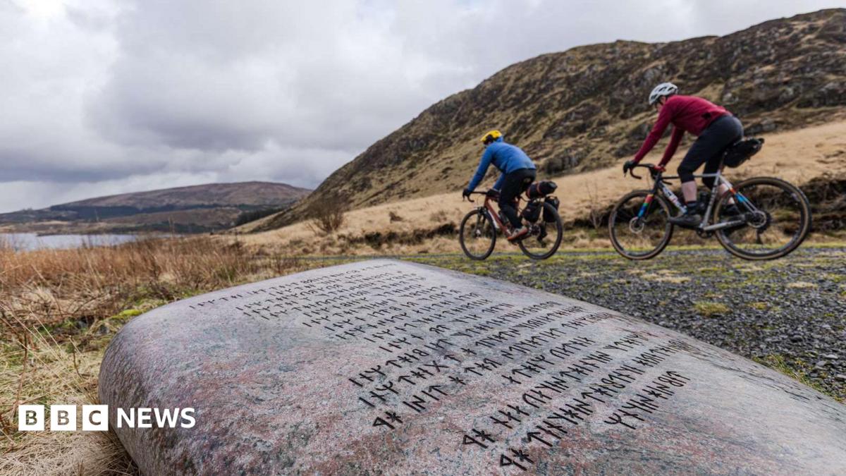 Two people on bikes cycle past a large stone with rugged hills in the background and a loch up ahead