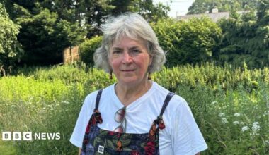 A woman with short grey hair is wearing a white t-shirt and dungarees, which have flowers on them. She is standing in front of wasit-high green plants and a few wildflowers. She has green framed sunglasses hanging from her t-shirt.