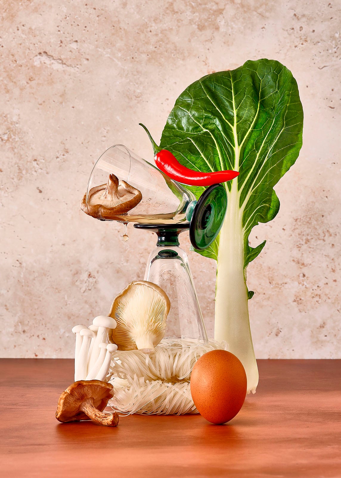 A creative arrangement of mushrooms, a red chili, bok choy, an egg, and glassware, with some ingredients balanced atop overturned glasses against a beige textured background.