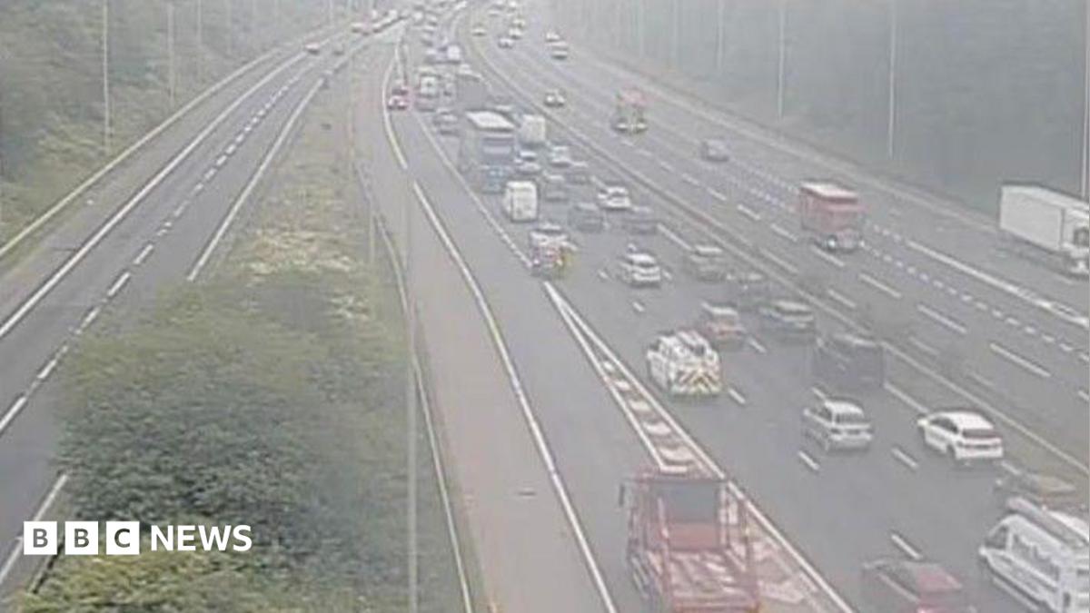 A traffic cam showing a motorway at a junction. The road is lined with thick woodland. On the left, a slip road joins the main motorway which is heavy with traffic moving away from the camera. The other side of the central reservation contains lighter traffic and a white lorry is leaving via a slip road which disappears from view.