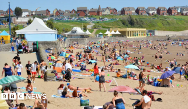 People on Barry Island beach in the sunshine. The beach is packed, with crowds lying on the sand, with the promenade and a row of houses in the background.
