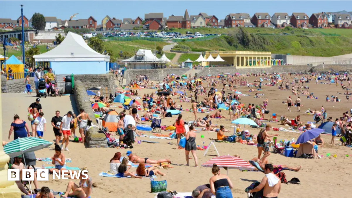 People on Barry Island beach in the sunshine. The beach is packed, with crowds lying on the sand, with the promenade and a row of houses in the background.