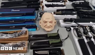 A table is filled with various knives and machetes sitting on top of their respective boxes. Some are wrapped in plastic packaging. In the centre of the image is a whole-head mask of a bald person.