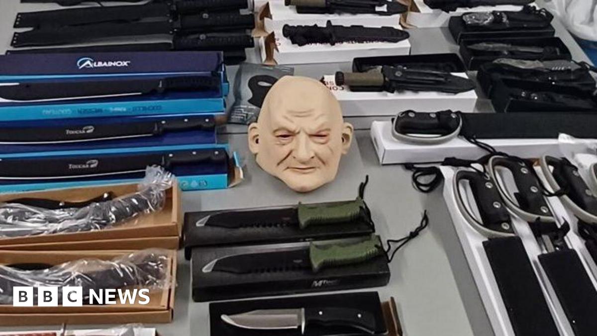 A table is filled with various knives and machetes sitting on top of their respective boxes. Some are wrapped in plastic packaging. In the centre of the image is a whole-head mask of a bald person.