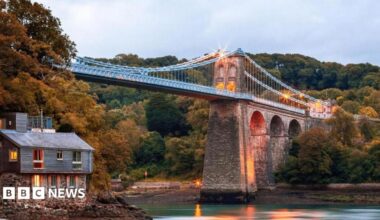 Menai Suspension Bridge, Anglesey, pictured from The Belgian Promenade. A house can be seen on the left hand side under the bridge. It is dawn and lights illuminate the bridge.