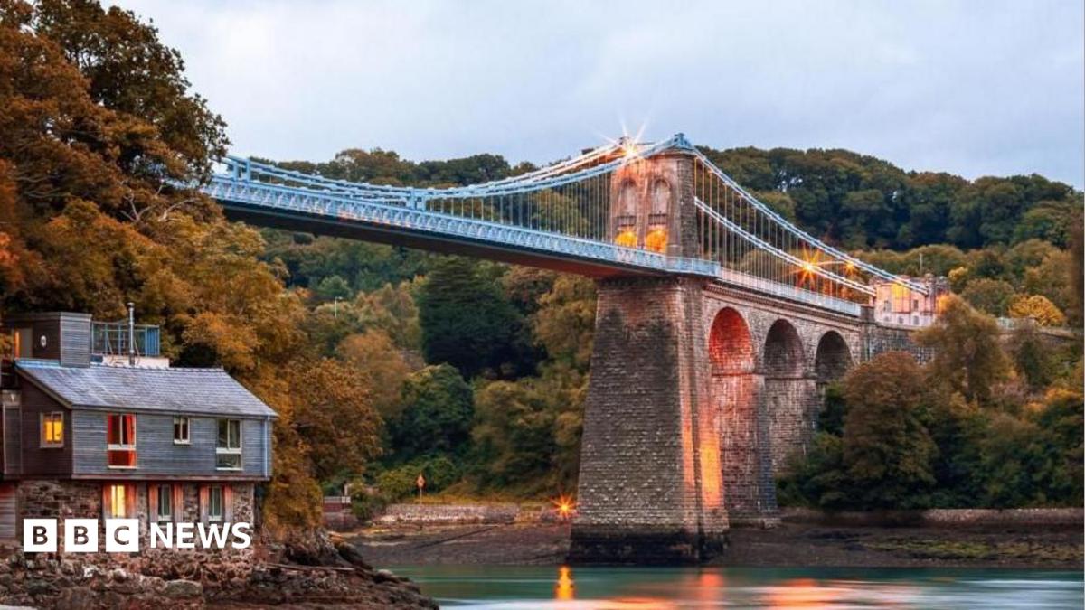 Menai Suspension Bridge, Anglesey, pictured from The Belgian Promenade. A house can be seen on the left hand side under the bridge. It is dawn and lights illuminate the bridge.