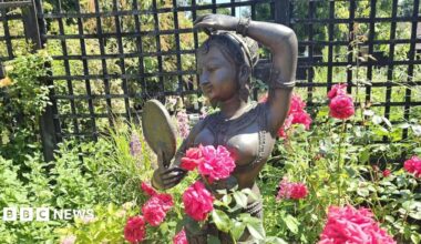 A bronze statue of an Indian woman is placed among pink flowers in a garden. She is designed as if wearing ornate clothing and looking at herself in a mirror. There are shrubs behind her and a trellis fence.