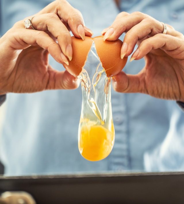 A detail of cracked egg falling into the pan as woman holds egg shells in both hands.