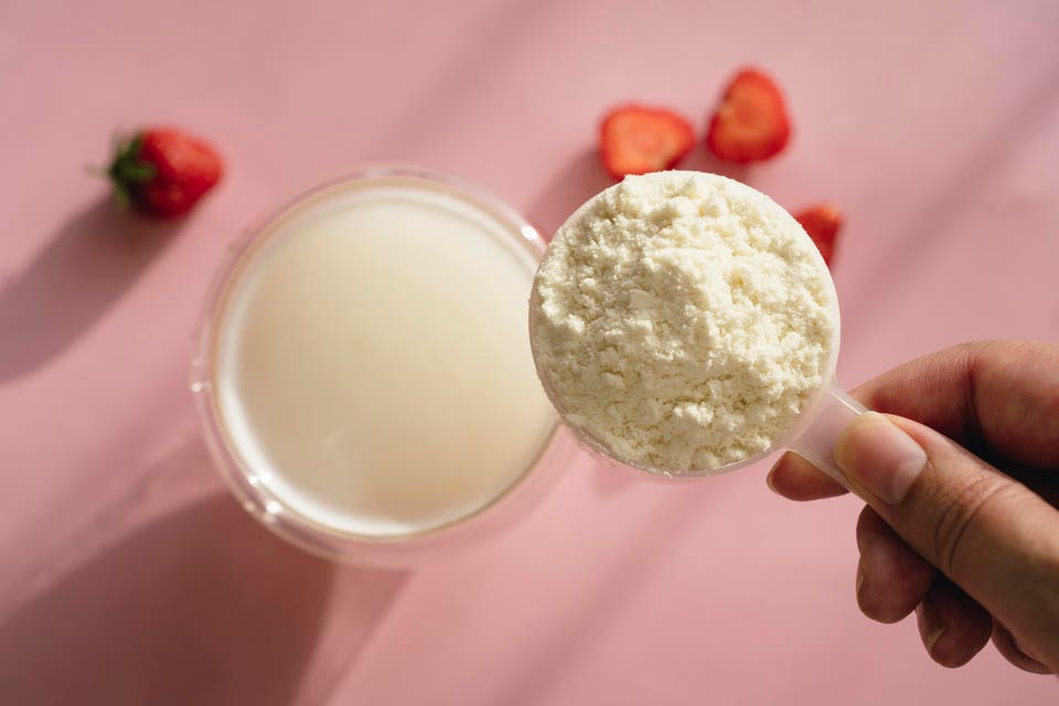 Whey protein powder in a measuring scoop, protein shake in a glass and strawberry on pink table in sunlight