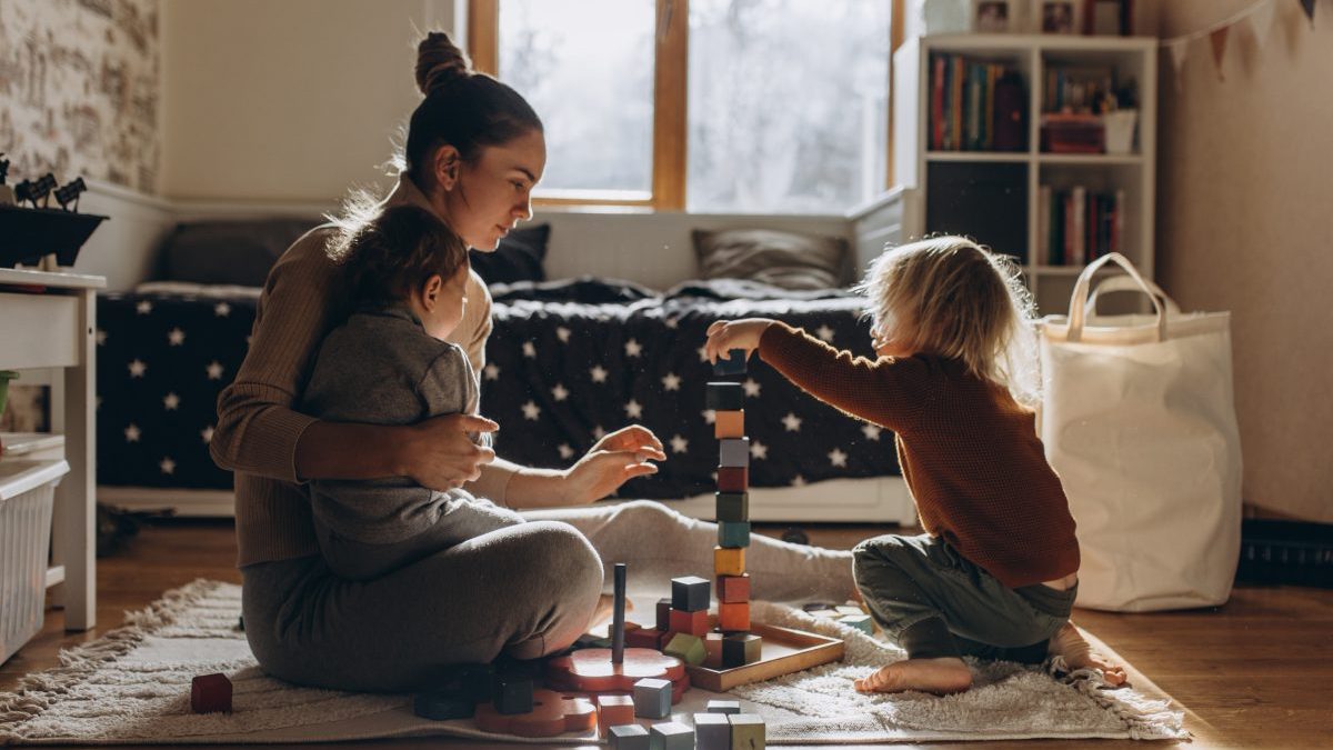 Young Mother playing with children while sitting on floor at home with wooden toys - stock photo