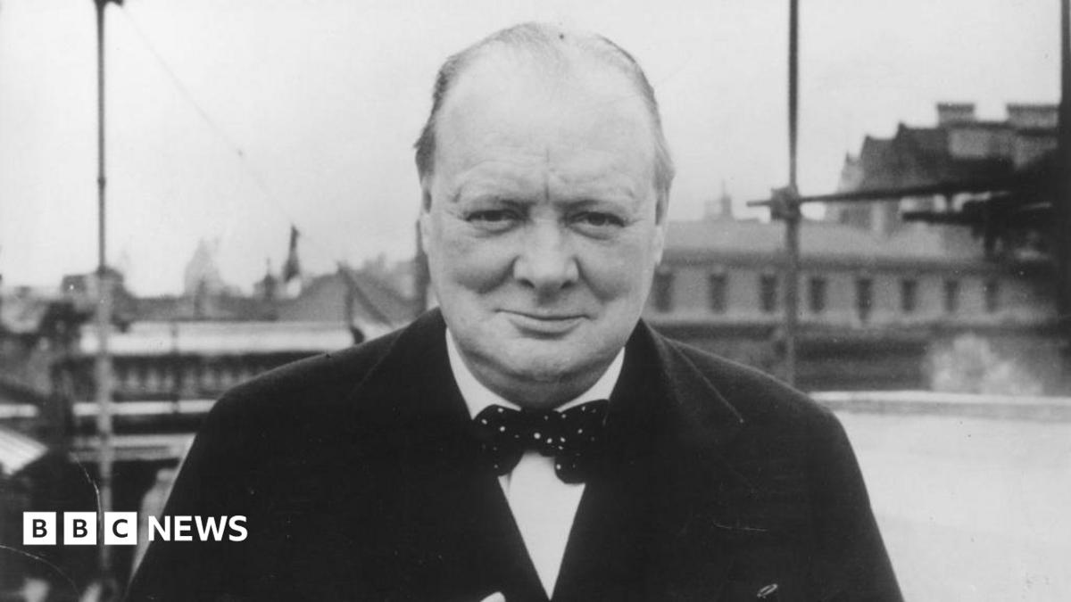 A black and white picture of Winston Churchill. He is looking directly into the camera. He is wearing a dark jacket, light shirt and a spotted bow tie.