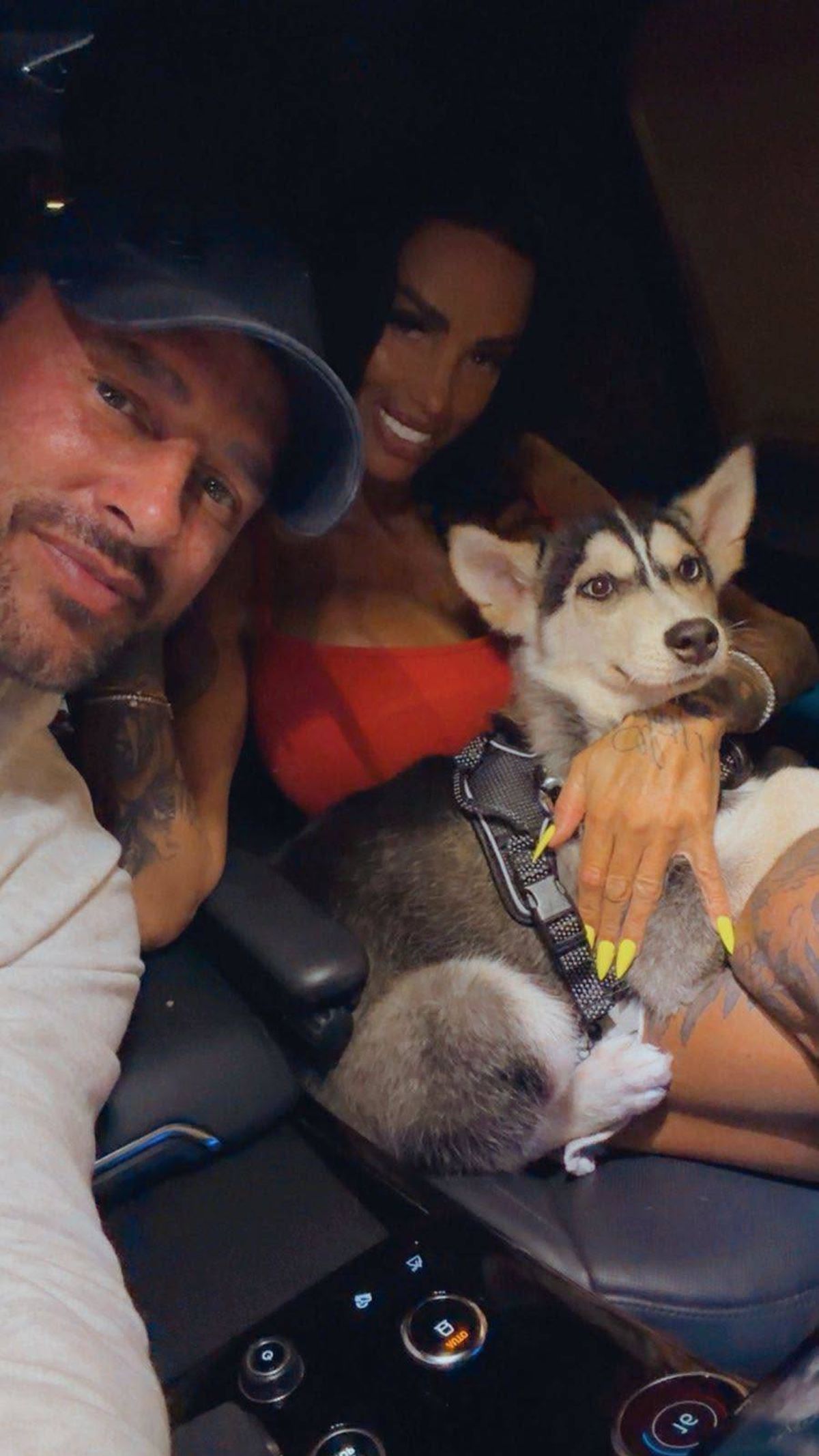 An adult male and female, both wearing casual attire, are smiling while posing for a photograph. The male individual dons a cap, while the female individual wears a red top. They are accompanied by a husky dog that appears to be the focal point of their attention.