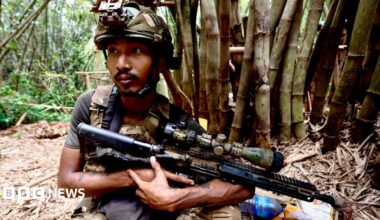 A soldier in a camouflage helmet holds a gun in a jungle setting, a tree trunk behind him. He has a watchful expression and his eyes are fixed beyond the camera.