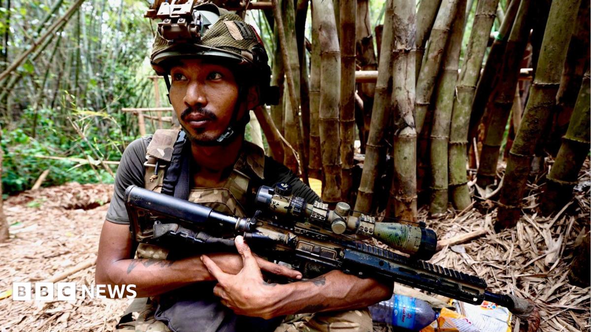 A soldier in a camouflage helmet holds a gun in a jungle setting, a tree trunk behind him. He has a watchful expression and his eyes are fixed beyond the camera.