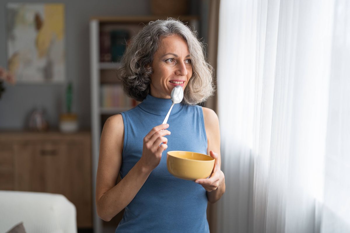 Mature woman with gray hair smiling while eating breakfast by a window