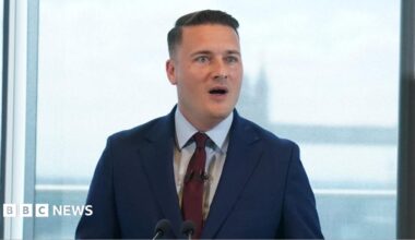 Wes Streeting wearing a navy suit and a red tie. He is standing at a lectern on front of a view of Tower Bridge in central London