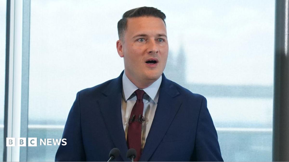 Wes Streeting wearing a navy suit and a red tie. He is standing at a lectern on front of a view of Tower Bridge in central London