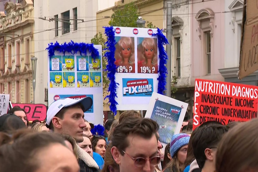 A large group of people stand on a city road holding up signs including one that says "fix allied health".