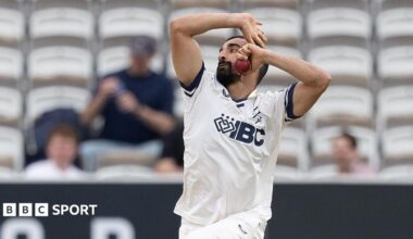Zafar Gohar about to release the ball as he bowls for Middlesex, with white seats and a few spectators in the background